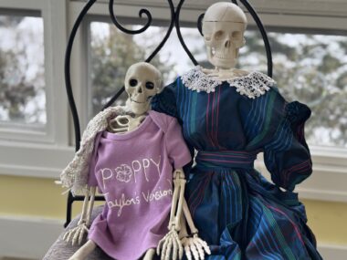 Two skeletons sit in a black iron chair in what appears to be a house. The one at left wears a lavender shirt that reads "Poppy Taylor’s version," with a a small crocheted scarf over one shoulder. The taller skeleton on the right wears a vintage blue plaid dress with a lace collar.