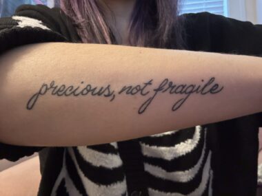 A forearm tattoo reads, in cursive, "precious, not fragile." It belongs to a woman in a black sweater with skeleton ribs down the front; we only see her torso. 