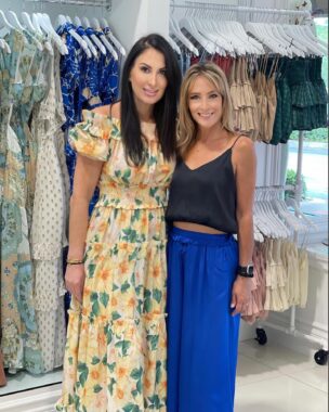 Two women stand next to each other embracing in front of racked dresses in a beach-themed boutique clothing store.