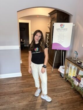 A young woman wearing a black shirt and light slacks smiles for a photo at the chiropractic office where she works.