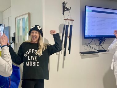 A woman with a shirt that says "Support Live Music" raises her left fist in triumph after ringing a bell, which actually looks like a wind chime, in a clinic hallway.