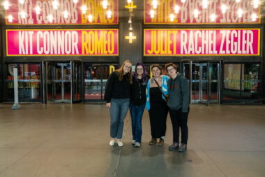 Four adults stand side by side in front of a theater entrance. Above the doors are pink lit-up signs that read "Romeo + Juliet" in the center with the actors' names, Kit Connor and Rachel Zegler, on either side.