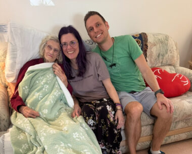 Three people sit on a couch: an elderly woman, a younger woman, and a man.