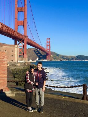 A mother and son stand at the base of the Golden Gate Bridge on a sunny day with clear, blue skies.