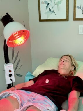 A woman with blond hair, a dark shirt, and shorts lies with her eyes closed on a table, with a red lamp aimed at her midsection from her right. The walls are gray, with a couple of flower prints visible behind the woman.