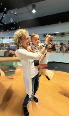 A woman, perhaps in her 50s, laughs while holding up a baby. They are inside a large room with a wooden floor, and there are bubbles in the air behind them.