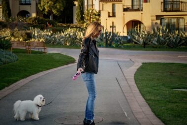 A woman walks a small, white dog on a leash on a sunny day. 