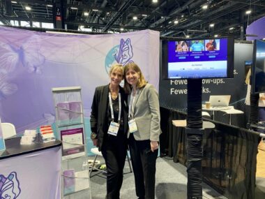 Two women stand close together for a photo pose in front of a purple background with some type of electronic screen in front of them. They're both wearing lanyards indicating they're attending some type of conference together. 