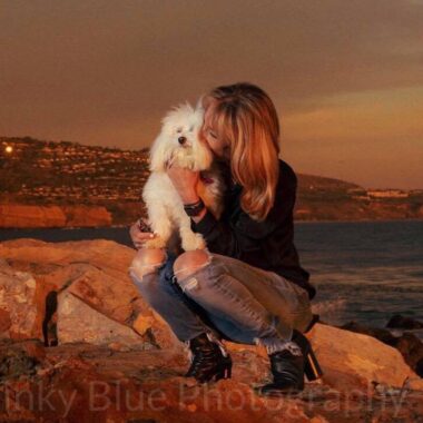A woman kneels atop rocks by the shoreline and snuggles her small dog.