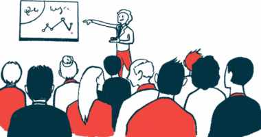 A person in red pants points to a graph as a large, seated audience watches.
