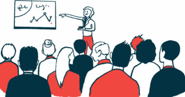 A person in red pants points to a graph as a large, seated audience watches.
