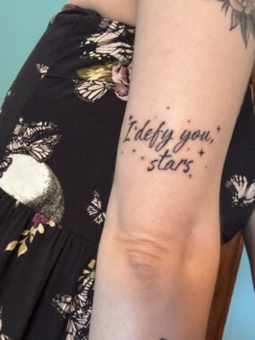 A photo of a woman's arm with a tattoo that reads "I defy you, stars."