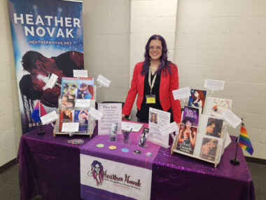 A woman with purple hair and glasses stands behind a table wearing a black shirt, red blazer, and lanyard. There's a large banner behind her and a smaller sign on her table featuring her name, and several of the books she's written are propped up on the table.
