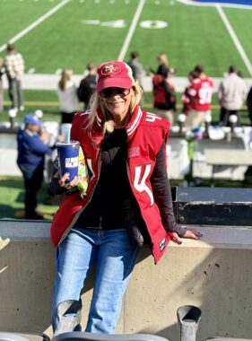 A woman rests against a wall in front of a football field. She's wearing a red baseball cap and vest with the San Francisco 49ers logo.