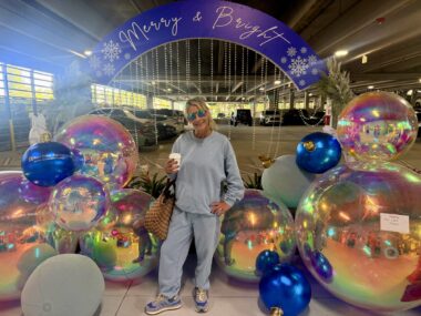 A woman in gray sweatpants and sweatshirt and mirrored sunglasses, and holding a cup of coffee, poses among shiny, colorful, metallic-looking large balloons and a purple banner that says "Merry and Bright." 