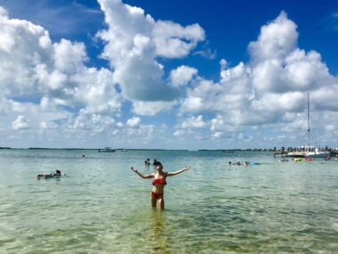 A woman stands knee-deep in the ocean with her arms outstretched. The water is clear and a beautiful shade of turquoise.