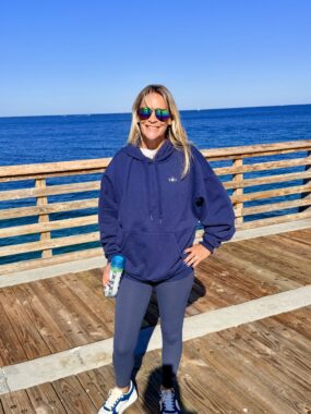 A woman in a blue jacket and sunglasses stands on a pier in front of a blue body of water and blue skies. 