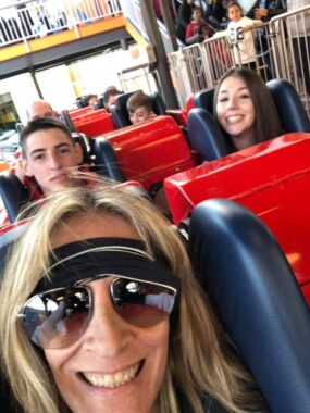 A woman takes a selfie in her seat on a roller coaster. Her daughter and son also smile at the camera from the row behind her.