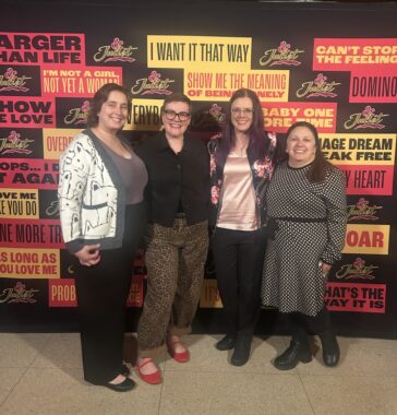 Four women stand together in front of a theater display.