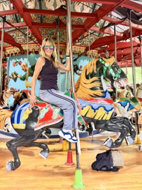 A woman in mirrored sunglasses and warm-weather clothing sits on a colorful carousel horse and smiles. 