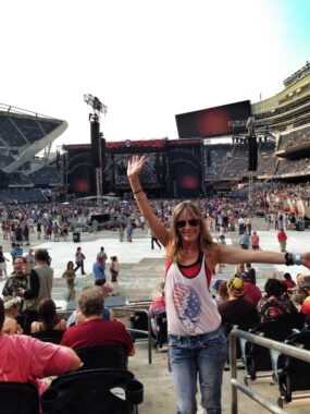 A woman wearing a Grateful Dead tank top stands in a stadium with her arms outstretched before a concert.