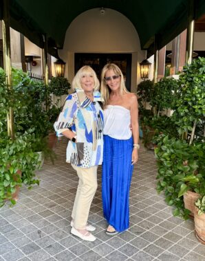 Two women - a mother and daughter - pose in the entryway to what looks like a fancy restaurant or club. They're both wearing light-colored outfits whose blue, white, and beige color schemes complement one another. 