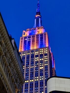 A photo from a phone shows the top of the Empire State Building, which is lit up with orange and blue lights. The blue lights are the same tone as the night sky.