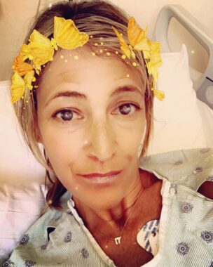A woman takes a selfie from her hospital bed. She's wearing a hospital gown and has at least one lead attached to her chest. The photo has a filter that makes it look like golden butterflies are encircling the woman's head.