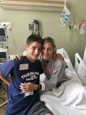 A teenage boy and his mom embrace in a hospital room. The mom is in a hospital bed.
