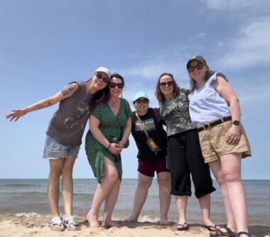 Five women pose on the shore of Lake Michigan in summer clothing on a sunny day. 