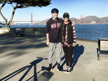 A woman and her teenage son pose for a photo in front of the Golden Gate Bridge. The son is wearing a bike helmet and has his arm around his mom's shoulders. 